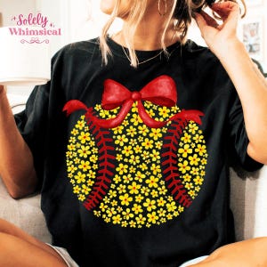 May include: Black t-shirt featuring a baseball design made of yellow flowers and red stitching. A red bow sits atop the floral baseball. The text "Solely Whimsical" is in the upper left corner.