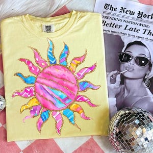 May include: A yellow Comfort Colors t-shirt featuring a colorful sun design. The sun has pink, blue, and yellow rays with gold glitter accents. A newspaper with the headline "Better Late Than" is in the background.
