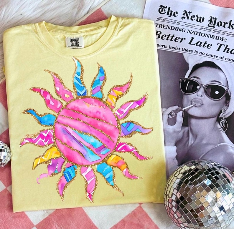 May include: A yellow Comfort Colors t-shirt featuring a colorful sun design. The sun has pink, blue, and yellow rays with gold glitter accents. A newspaper with the headline "Better Late Than" is in the background.
