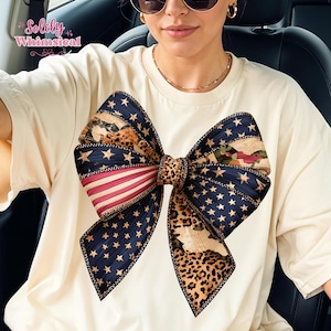 Op de afbeelding: Crèmekleurig T-shirt met een groot strikdesign. De strik bevat elementen van de Amerikaanse vlag met rode en witte strepen, blauwe sterren en een luipaardprint. De tekst "Solely Whimsical" staat in de linkerbovenhoek.