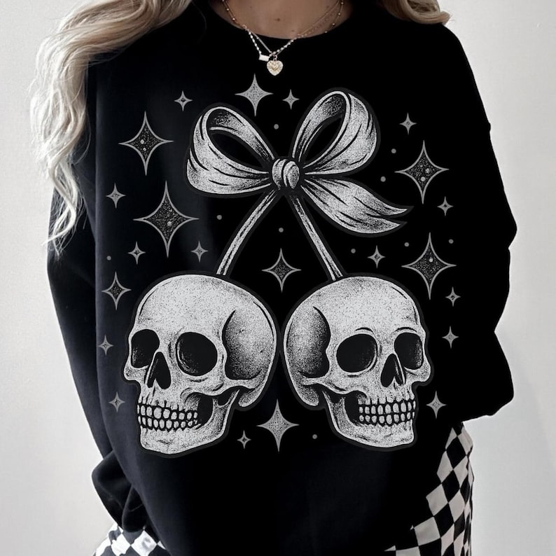K&ouml;nnte beinhalten: Schwarzes Sweatshirt mit einem wei&szlig;en Motiv aus zwei Sch&auml;deln, die durch eine Schleife verbunden sind, die an Kirschen erinnert. Das Design umfasst Sterne. Ein l&auml;ssiges Kleidungsst&uuml;ck, geeignet f&uuml;r den Alltag.