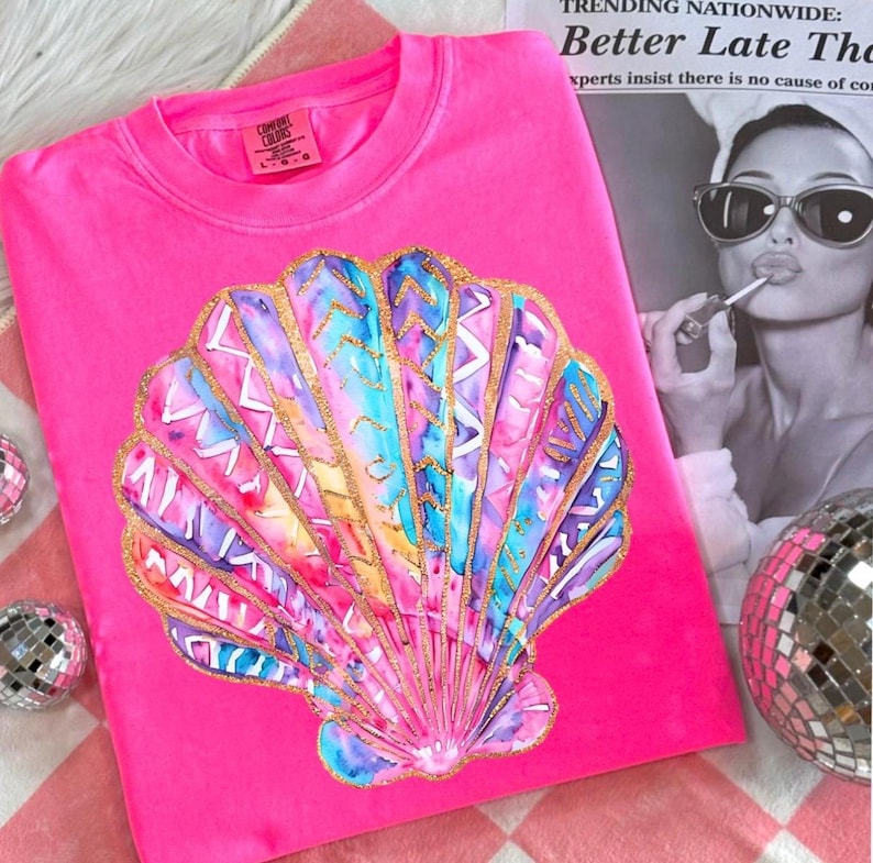 May include: A bright pink sweatshirt featuring a large, colorful seashell design. The seashell is painted with watercolor-style hues of blue, purple, and gold. The text "Comfort Colors" is visible on the label. The background includes a black and white image and disco balls.