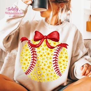 May include: Cream-colored sweatshirt featuring a softball design made of yellow flowers, red stitching, and a red bow. The text "Solely Whimsical" is printed in the upper left corner. A casual, comfortable top for everyday wear.