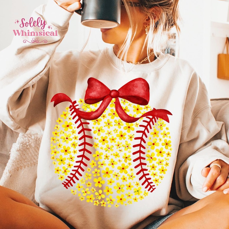 May include: Cream-colored sweatshirt featuring a softball design made of yellow flowers, red stitching, and a red bow. The text "Solely Whimsical" is printed in the upper left corner. A casual, comfortable top for everyday wear.