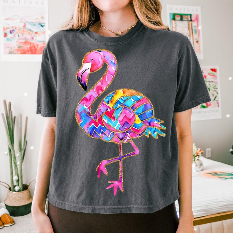 May include: Dark gray t-shirt featuring a colorful flamingo design. The flamingo has a pink head and legs, a yellow beak, and a body with a vibrant pattern of blue, yellow, and purple zigzags. The design is outlined in gold glitter.