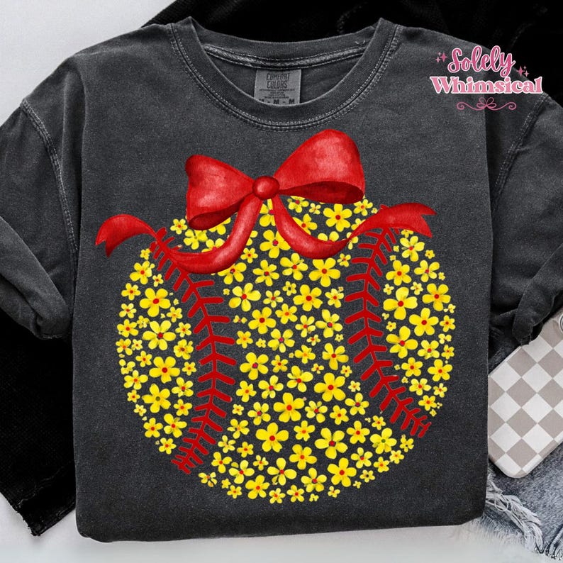 May include: Dark gray sweatshirt featuring a baseball design made of yellow flowers and red stitching. A red bow adorns the top of the baseball. The text "Solely Whimsical" is in the upper right corner.