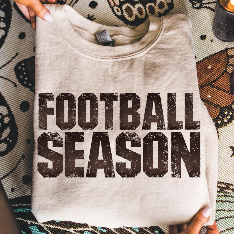 May include: A beige sweatshirt featuring the text "FOOTBALL SEASON" in a distressed, dark brown font. The folded sweatshirt is held by hands, with a patterned fabric visible in the background.