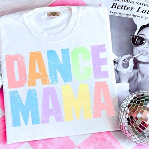 May include: White t-shirt with the words "DANCE MAMA" in colorful, distressed lettering. The shirt has a crew neck and a tag at the neckline. A disco ball and a black and white image are in the background.