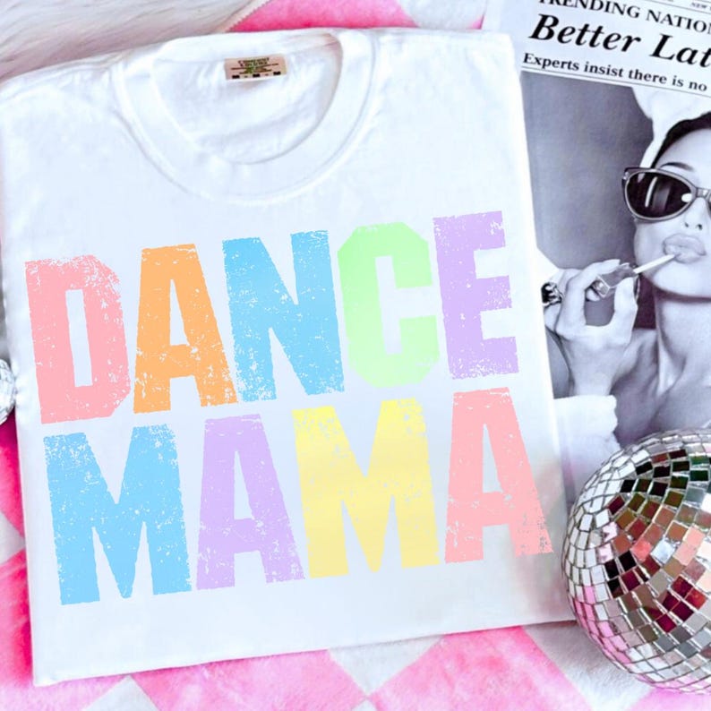 May include: White t-shirt with the words "DANCE MAMA" in colorful, distressed lettering. The shirt has a crew neck and a tag at the neckline. A disco ball and a black and white image are in the background.