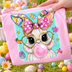May include: Pink t-shirt featuring a cartoon bunny with heart-shaped glasses blowing a bubble. The bunny has a pink bow with white polka dots and rainbow ears. The shirt has the text "Solely Whimsical".