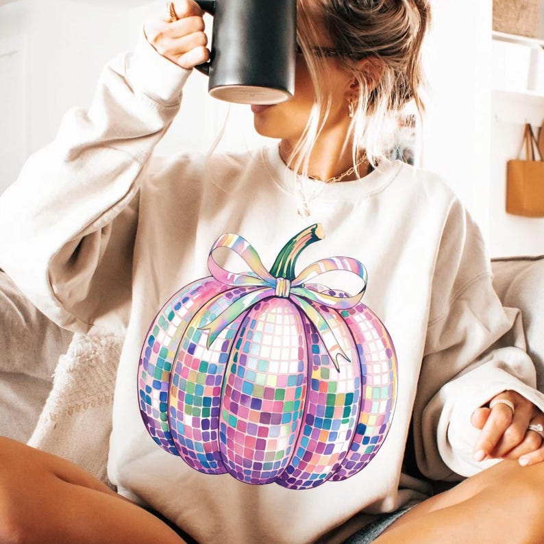 Puede incluir: Una sudadera color crema con un dise&ntilde;o de calabaza colorido estilo mosaico. La calabaza tiene un tallo verde y un gran lazo iridiscente. El dise&ntilde;o est&aacute; formado por peque&ntilde;as baldosas cuadradas en tonos rosa, morado, azul y amarillo.