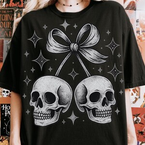 K&ouml;nnte beinhalten: Schwarzes T-Shirt mit einer Grafik von zwei Sch&auml;deln, die durch eine Schleife verbunden sind, mit Sternakzenten. Die Sch&auml;del sind schattiert und die Schleife ist hellgrau. Das Design hat ein gotisches oder Halloween-Thema.
