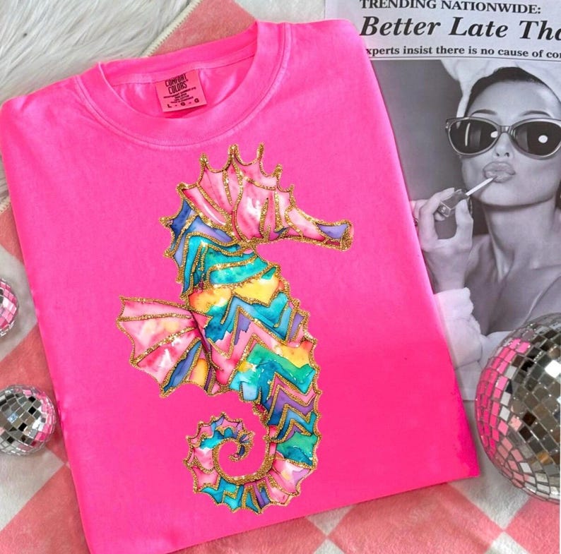 May include: A bright pink sweatshirt featuring a colorful seahorse design. The seahorse is decorated with pink, blue, yellow, and gold glitter accents. The sweatshirt is a Comfort Colors brand.