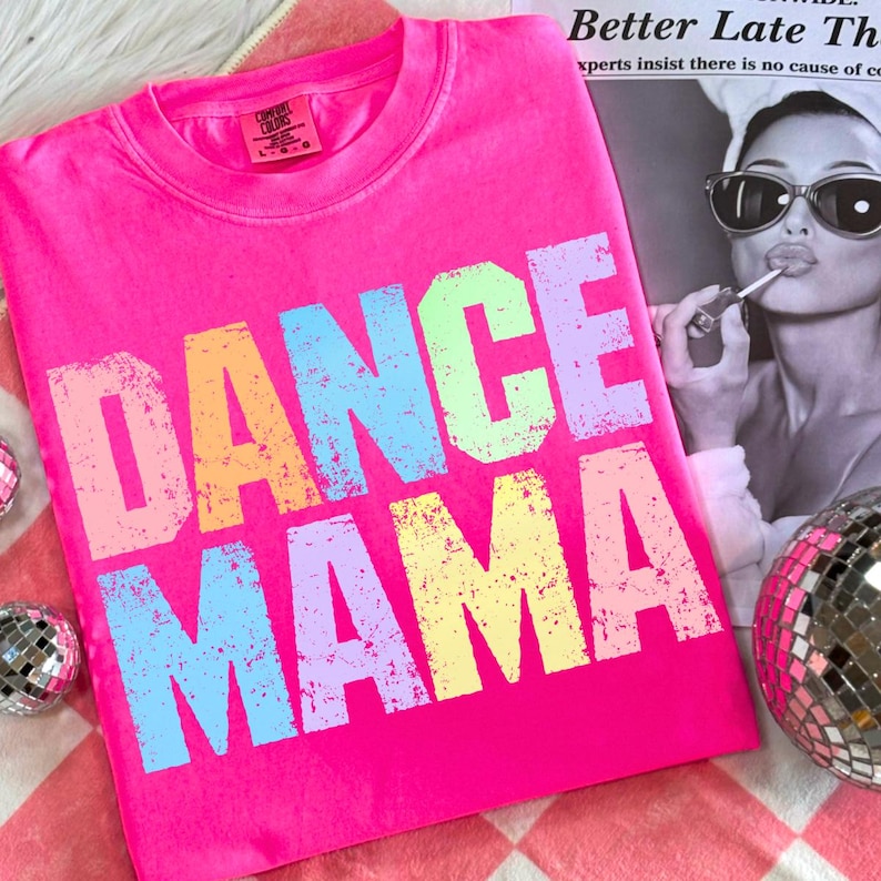May include: A hot pink t-shirt with the words "DANCE MAMA" printed in large, distressed, pastel-colored letters. The shirt is folded and lies on a red and white checkered surface. A disco ball and a black and white magazine are in the background.