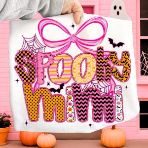 Peut inclure: Une couverture blanche sur le th&egrave;me d'Halloween avec les mots "Spooky Mimi" dans une police color&eacute;e et &agrave; motifs. Le motif comprend un n&oelig;ud rose, des toiles d'araign&eacute;es, des chauves-souris et des citrouilles. La couverture est tenue devant un fond rose avec des citrouilles.