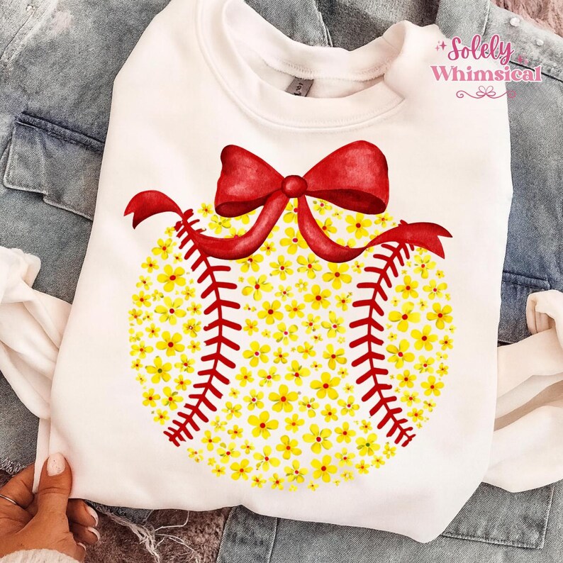 May include: White sweatshirt featuring a baseball design made of yellow flowers and red stitching. A red bow is at the top of the baseball design. The text "Solely Whimsical" is in the upper right corner. The sweatshirt is on a denim jacket.