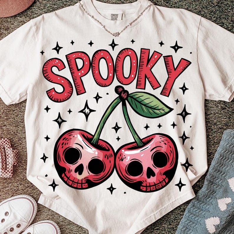 Może przedstawiać: Biała koszulka z upiornym wzorem wiśni. Słowo "SPOOKY" jest w czerwonych blokowych literach nad dwiema wiśniami z twarzami czaszek. Zielone łodygi i liście łączą wiśnie. Czarne gwiazdki otaczają wz&oacute;r.