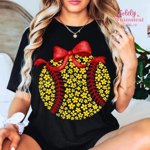 May include: Black t-shirt featuring a baseball design made of yellow flowers and red stitching, topped with a red bow. The shirt has short sleeves and a round neckline. The text "Solely Whimsical" is in the upper right corner.