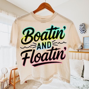 K&ouml;nnte beinhalten: Ein beiges T-Shirt mit den Worten "Boatin' and Floatin'" in einem schwarzen, t&uuml;rkisfarbenen, gelben und rosa Design. Das Shirt h&auml;ngt an einem Holzb&uuml;gel.