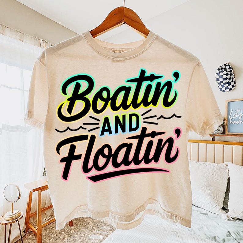 K&ouml;nnte beinhalten: Ein beiges T-Shirt mit den Worten "Boatin' and Floatin'" in einem schwarzen, t&uuml;rkisfarbenen, gelben und rosa Design. Das Shirt h&auml;ngt an einem Holzb&uuml;gel.