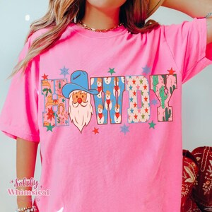 K&ouml;nnte beinhalten: Ein leuchtend pinkfarbenes T-Shirt mit dem Wort "COWBOY" in einem farbenfrohen, festlichen Design. Das "C" zeigt einen Weihnachtsmann mit Cowboyhut. Das "W" hat Schneem&auml;nner, und das "Y" hat Kakteen. Sterne und Schneeflocken sind um das Wort herum verstreut.