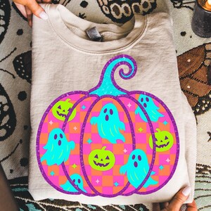 May include: A beige sweatshirt featuring a large, colorful Halloween-themed pumpkin design. The pumpkin has a teal stem, a pink and orange checkered body, and is decorated with teal ghosts and green jack-o'-lanterns. The design is outlined in purple.