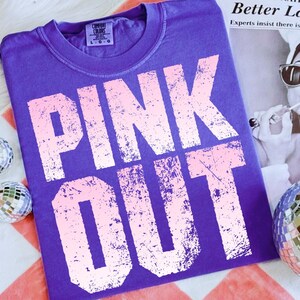 Puede incluir: Una camiseta morada Comfort Colors con las palabras "PINK OUT" en una fuente rosa claro desgastada. La camiseta est&aacute; colocada sobre una superficie a rayas rojas y blancas, con bolas de discoteca en el fondo.