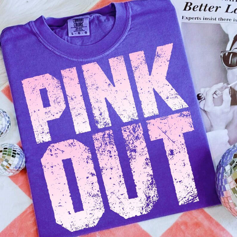 Puede incluir: Una camiseta morada Comfort Colors con las palabras "PINK OUT" en una fuente rosa claro desgastada. La camiseta est&aacute; colocada sobre una superficie a rayas rojas y blancas, con bolas de discoteca en el fondo.
