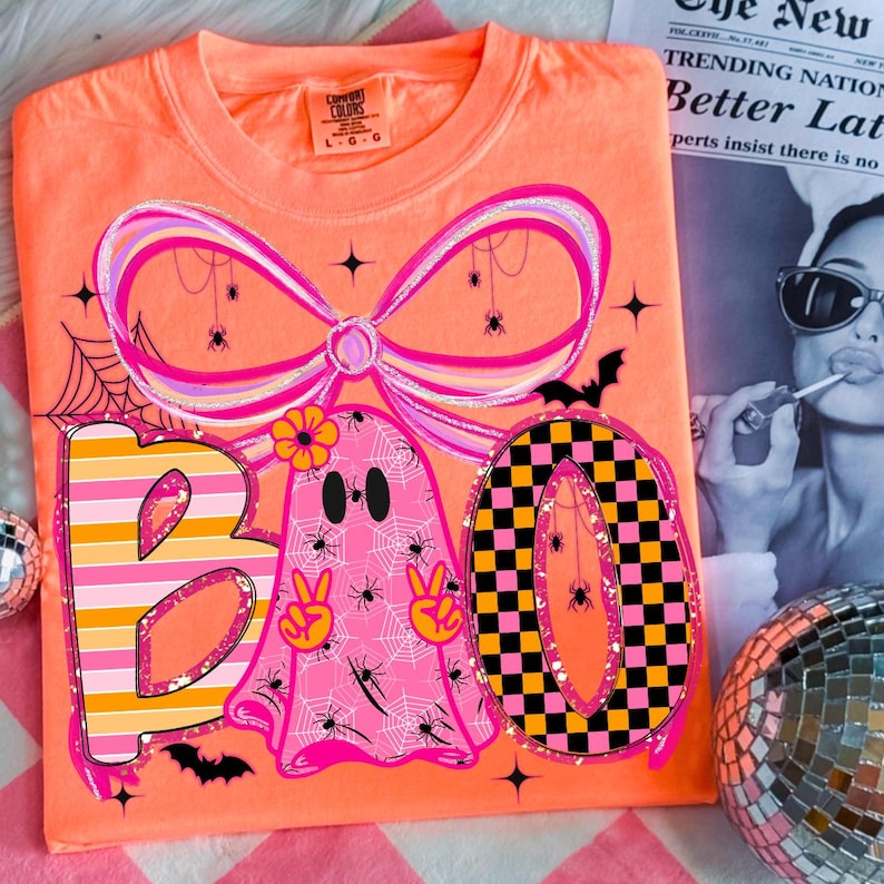 Op de afbeelding: Oranje Comfort Colors T-shirt met een Halloween-thema. Het ontwerp spelt "BOO" met een roze spook, een strik en een geblokte "O". De afbeelding bevat spinnen, spinnenwebben en vleermuizen.
