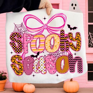 Pode incluir: Um cobertor branco com tema de Halloween com as palavras "Spooky Season" em uma fonte colorida e estampada. O design inclui morcegos, teias de aranha e um grande la&ccedil;o rosa. O cobertor &eacute; segurado em frente a um fundo rosa.