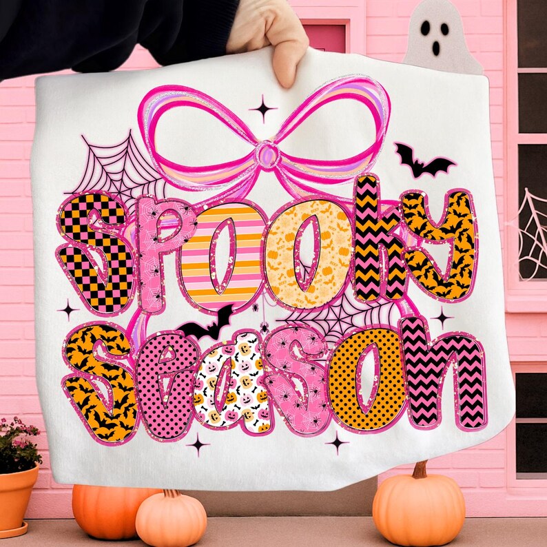 Pode incluir: Um cobertor branco com tema de Halloween com as palavras "Spooky Season" em uma fonte colorida e estampada. O design inclui morcegos, teias de aranha e um grande la&ccedil;o rosa. O cobertor &eacute; segurado em frente a um fundo rosa.