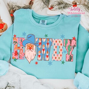 K&ouml;nnte beinhalten: Hellblaues Sweatshirt mit dem Wort "HOWDY" in einem farbenfrohen Muster. Das "H" zeigt einen Weihnachtsmann mit Cowboyhut. Die "O"s haben Schneem&auml;nner, und die "W" und "Y" haben Sterne und Kakteen.