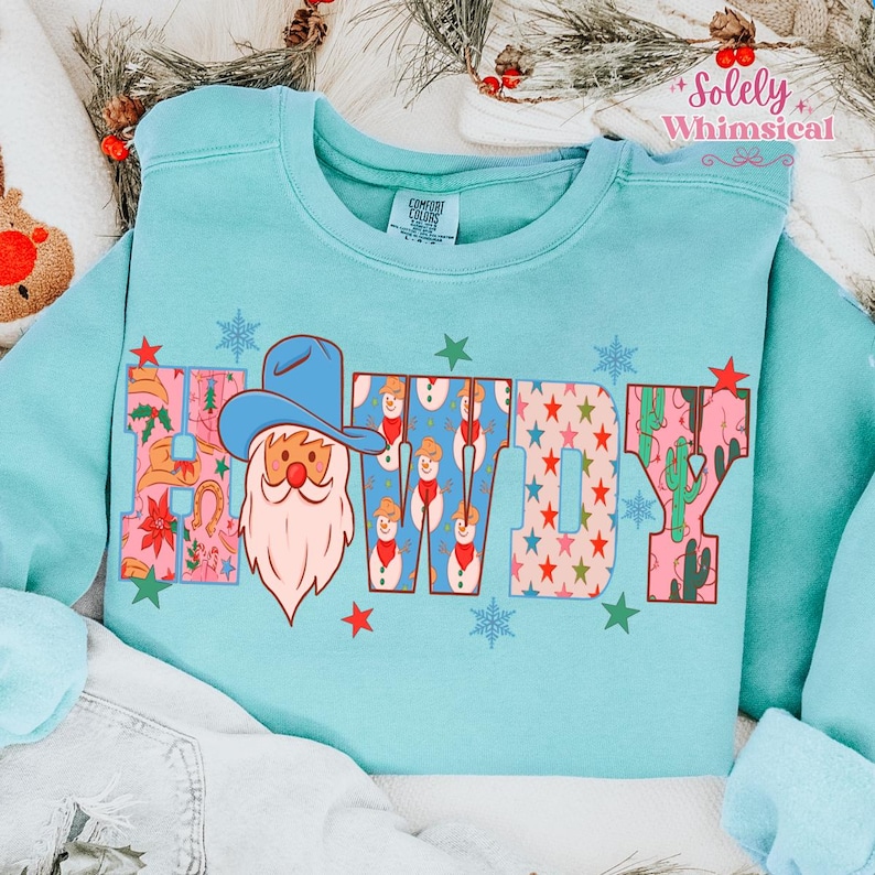 K&ouml;nnte beinhalten: Hellblaues Sweatshirt mit dem Wort "HOWDY" in einem farbenfrohen Muster. Das "H" zeigt einen Weihnachtsmann mit Cowboyhut. Die "O"s haben Schneem&auml;nner, und die "W" und "Y" haben Sterne und Kakteen.