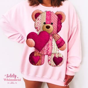 May include: A light pink sweatshirt featuring a patchwork teddy bear holding a heart. The bear is made of various pink and cream fabrics with heart and floral patterns. The sweatshirt has the text "Solely Whimsical" in the bottom left corner.