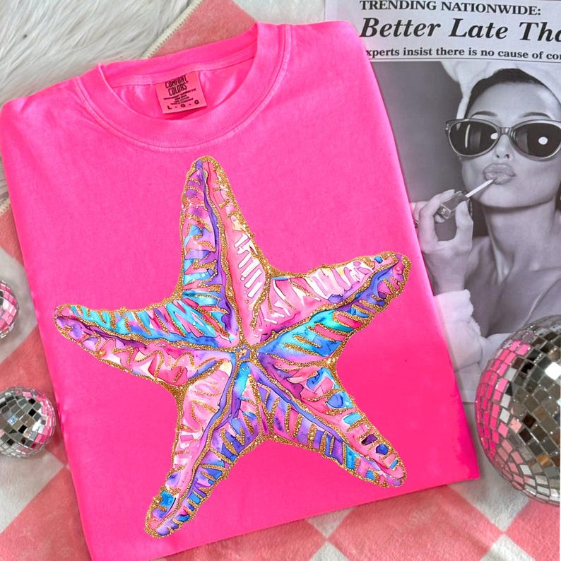 May include: Bright pink Comfort Colors sweatshirt featuring a large, colorful starfish design. The starfish is detailed with pink, blue, purple, and gold accents. The shirt is displayed on a pink and white checkered surface.