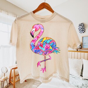 May include: A beige t-shirt featuring a colorful flamingo design. The flamingo has a pink head and legs, a black beak, and a body with a vibrant pattern of blue, purple, yellow, and orange zigzags. The shirt is hanging on a wooden hanger.
