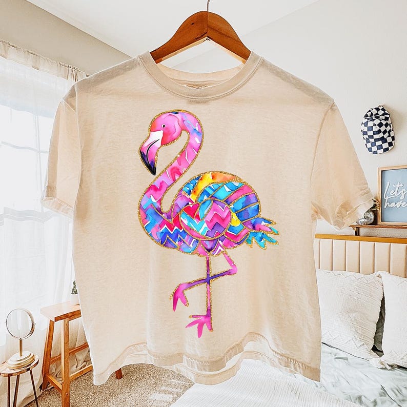 May include: A beige t-shirt featuring a colorful flamingo design. The flamingo has a pink head and legs, a black beak, and a body with a vibrant pattern of blue, purple, yellow, and orange zigzags. The shirt is hanging on a wooden hanger.