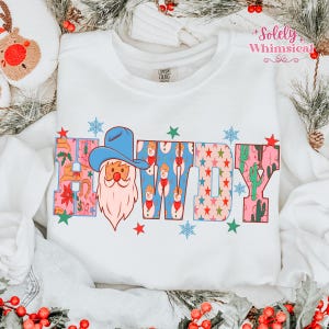 K&ouml;nnte beinhalten: Wei&szlig;es Sweatshirt mit dem Wort "Cowboy" in einem farbenfrohen Design. Das "C" zeigt einen Weihnachtsmann mit Cowboyhut, das "W" hat Schneem&auml;nner und das "Y" Kakteen. Das Sweatshirt hat den Text "Comfort Colors" auf der Vorderseite.