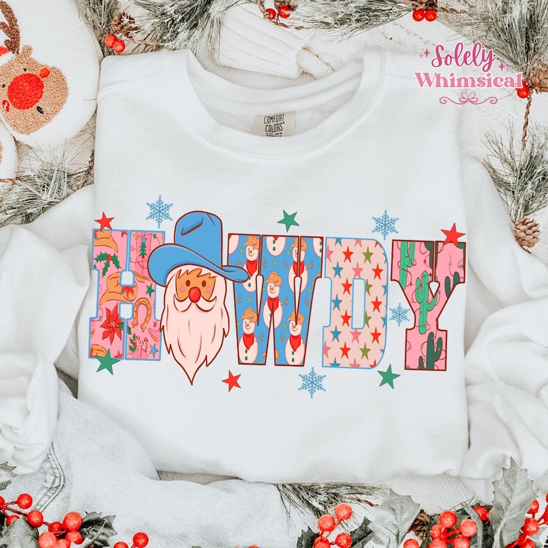 K&ouml;nnte beinhalten: Wei&szlig;es Sweatshirt mit dem Wort "Cowboy" in einem farbenfrohen Design. Das "C" zeigt einen Weihnachtsmann mit Cowboyhut, das "W" hat Schneem&auml;nner und das "Y" Kakteen. Das Sweatshirt hat den Text "Comfort Colors" auf der Vorderseite.
