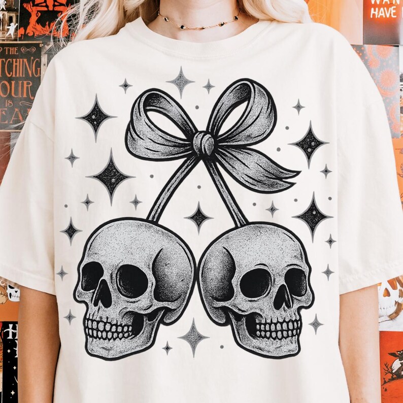 K&ouml;nnte beinhalten: Cremefarbenes T-Shirt mit einer schwarz-wei&szlig;en Grafik von zwei Sch&auml;deln, die durch eine Schleife verbunden sind und an Kirschen erinnern. Das Design enth&auml;lt Sterne. Ein l&auml;ssiges Kleidungsst&uuml;ck.