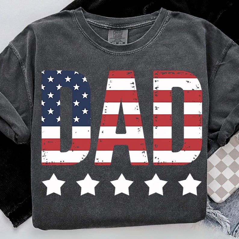 Puede incluir: Sudadera gris oscuro con la palabra "DAD" en rojo, blanco y azul, dise&ntilde;ada para parecerse a la bandera estadounidense. Debajo de la palabra "DAD" hay cinco estrellas blancas. Marca Comfort Colors.