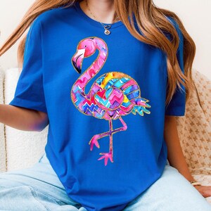 May include: A royal blue t-shirt featuring a colorful flamingo design. The flamingo has a pink head and legs, a black beak, and a body with a vibrant, multi-colored pattern. The design is outlined with gold glitter.