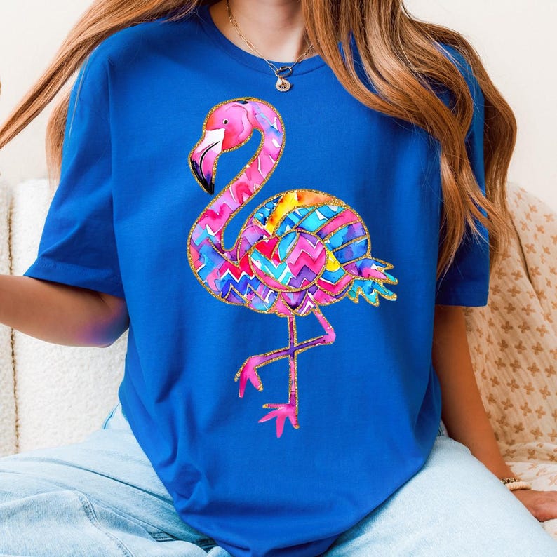 May include: A royal blue t-shirt featuring a colorful flamingo design. The flamingo has a pink head and legs, a black beak, and a body with a vibrant, multi-colored pattern. The design is outlined with gold glitter.