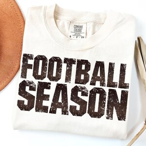 May include: Off-white t-shirt featuring the text "FOOTBALL SEASON" in a distressed, dark brown font. The shirt has a crew neck and a Comfort Colors label. A brown hat and other items are partially visible.