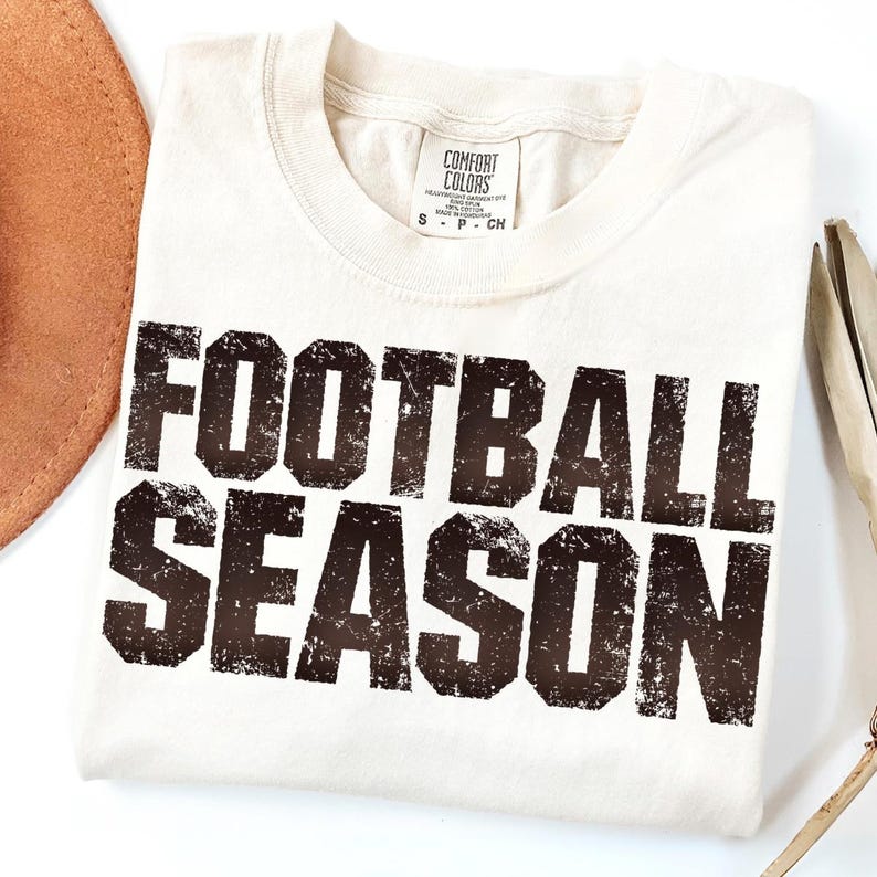 May include: Off-white t-shirt featuring the text "FOOTBALL SEASON" in a distressed, dark brown font. The shirt has a crew neck and a Comfort Colors label. A brown hat and other items are partially visible.