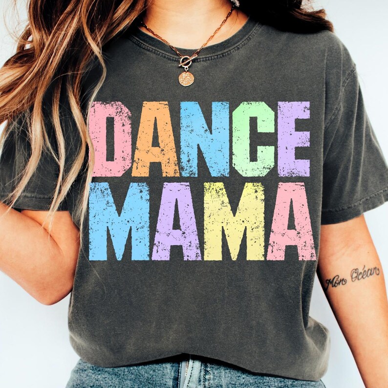 May include: Dark gray t-shirt with the words "DANCE MAMA" in large, colorful, distressed letters. The text is in pastel colors: pink, orange, blue, purple, and yellow. The shirt has short sleeves and a crew neck.