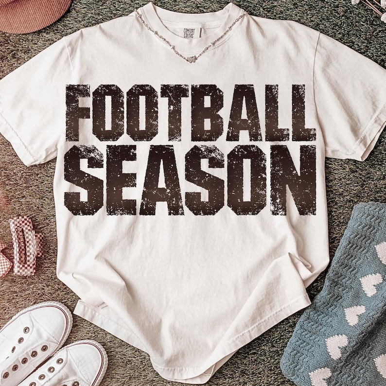 May include: A white t-shirt with the text "FOOTBALL SEASON" in a distressed, dark brown font. The shirt has a crew neck and short sleeves. Accessories include white trainers, a pink hat, and blue socks with white hearts.