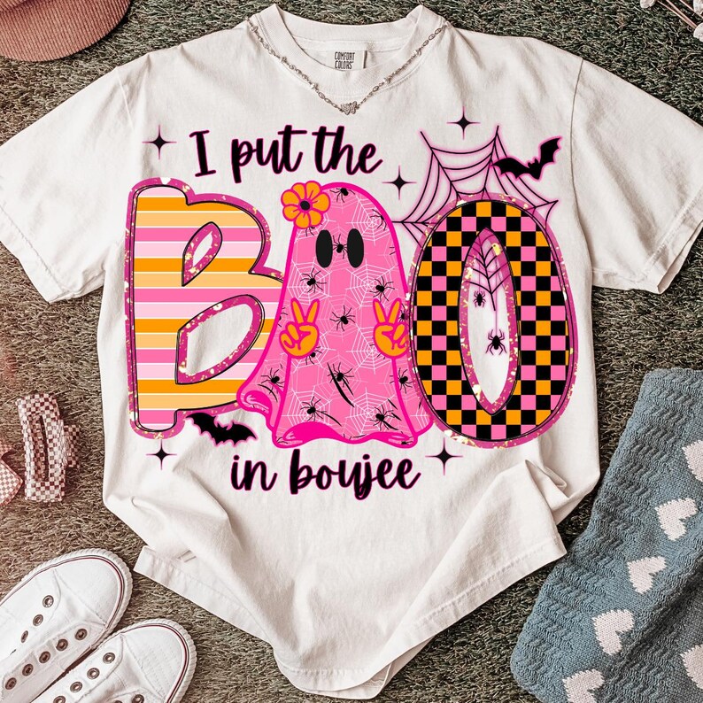 May include: White t-shirt with a Halloween-themed graphic. The design features the word "BOO" with a pink ghost, spiderwebs, and the phrase "I put the BOO in boujee". The letters are in pink, orange, and black.