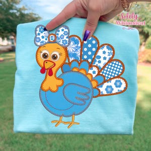 Könnte beinhalten: Ein hellblaues T-Shirt mit einem Cartoon-Truthahn-Design. Der Truthahn hat einen orangefarbenen Kopf und Beine, einen blauen Körper und eine Schleife mit floralem Muster. Die Schwanzfedern des Truthahns sind blau und weiß mit Blumen- und Tupfenmustern. Der Text "Solely Whimsical" befindet sich in der oberen rechten Ecke.