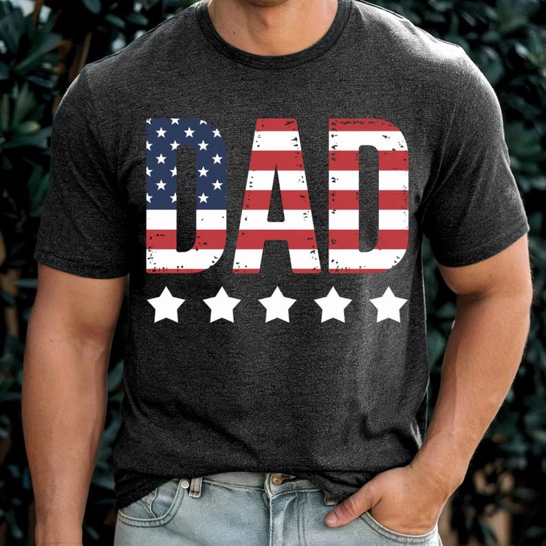Puede incluir: Camiseta gris oscuro con la palabra "DAD" en rayas rojas y blancas y estrellas azules, que recuerda a la bandera americana. Cinco estrellas blancas est&aacute;n debajo. La camiseta es de manga corta.